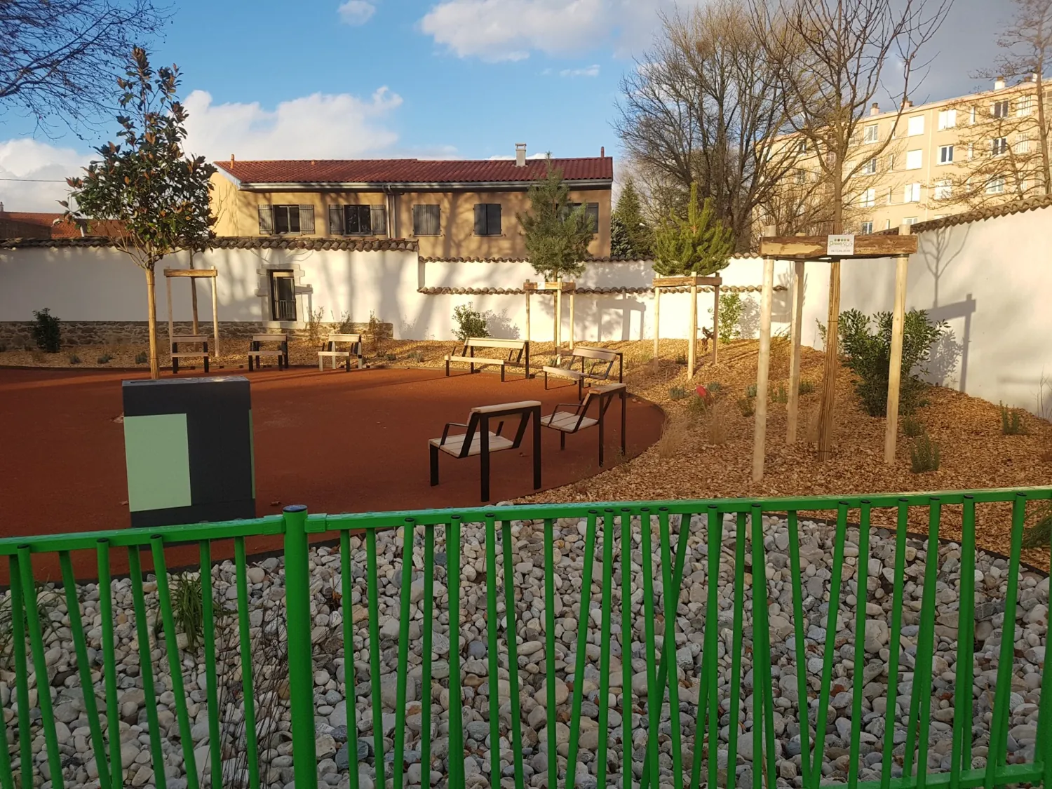 Petit parc urbain avec bancs, jeunes arbres tuteurés et massifs paillés derrière une clôture verte