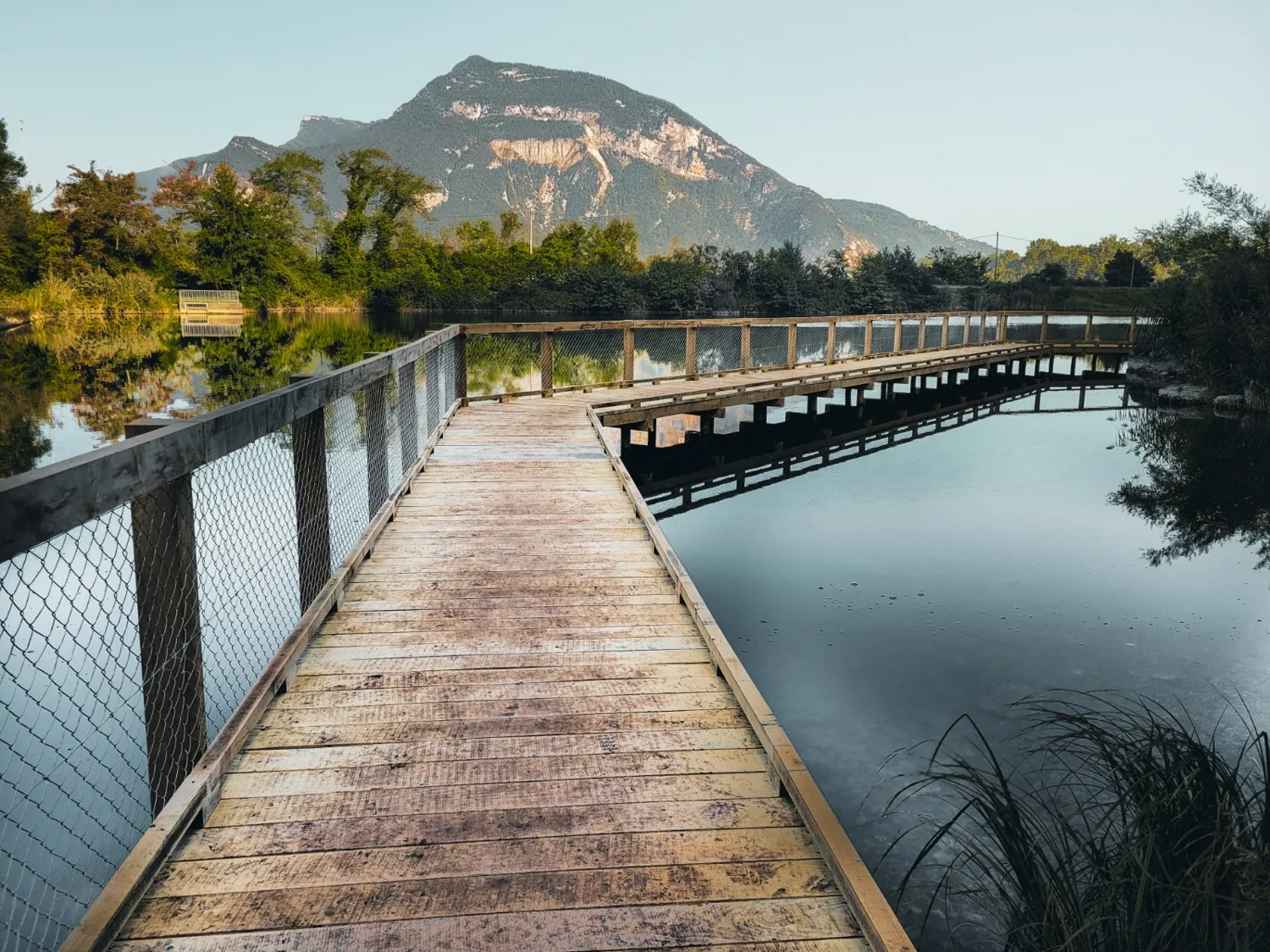 Passerelle en bois avec garde-corps au-dessus d'un plan d'eau, entourée de végétation au pied d'une montagne