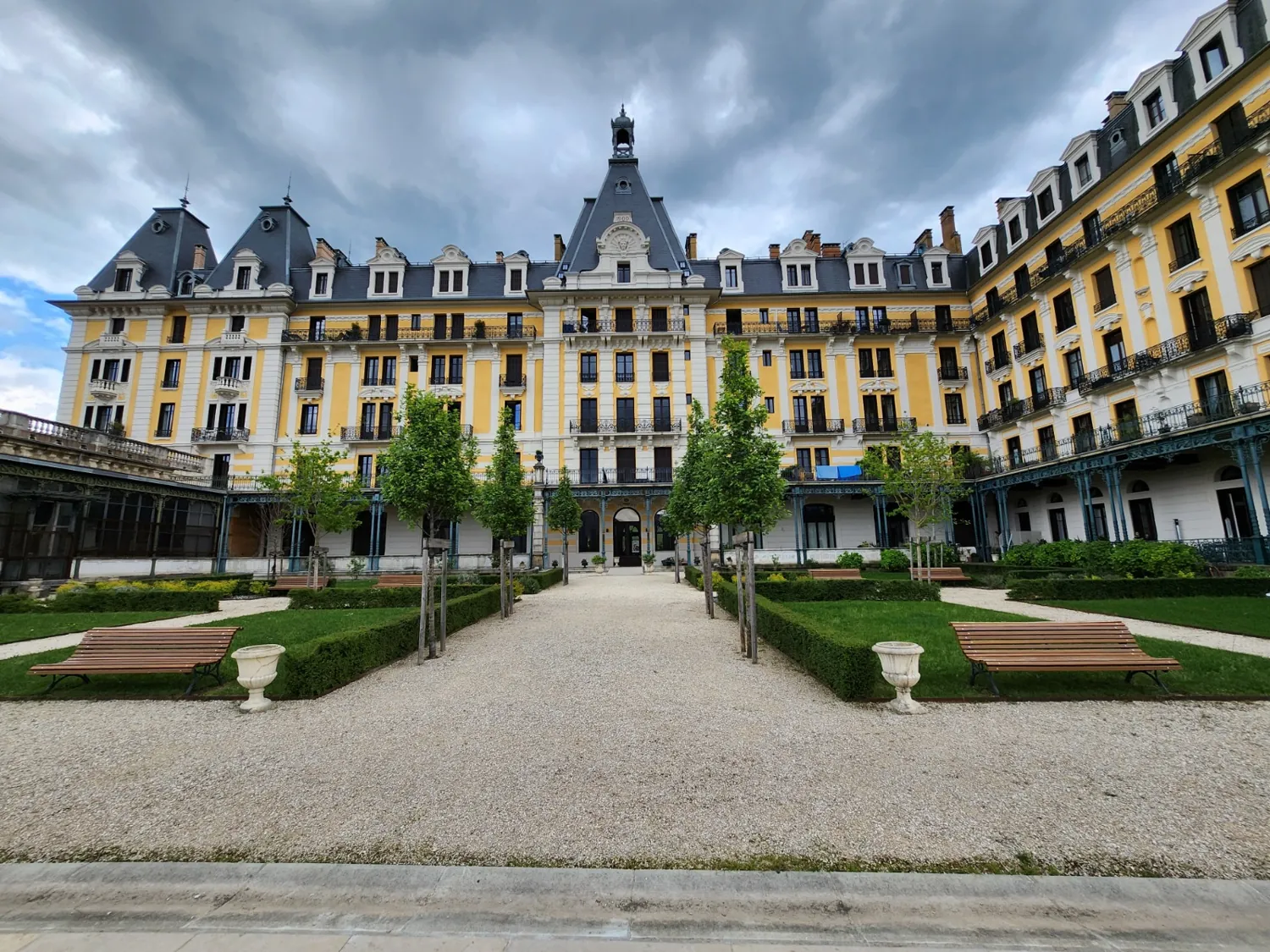 Allée de gravier bordée d'arbres, de haies taillées et de bancs devant un grand bâtiment à façade jaune