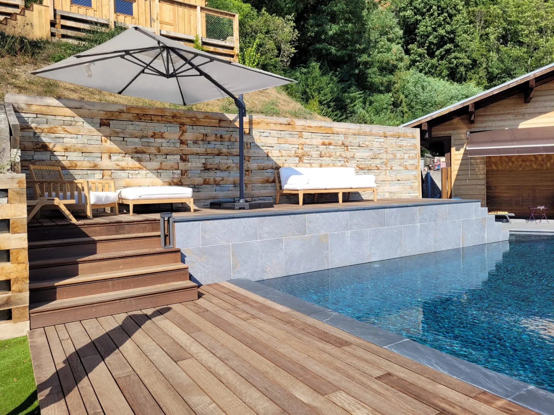 Terrasse en bois, mur en pierre et salon sous parasol au bord de la piscine – Veyrier-du-Lac
