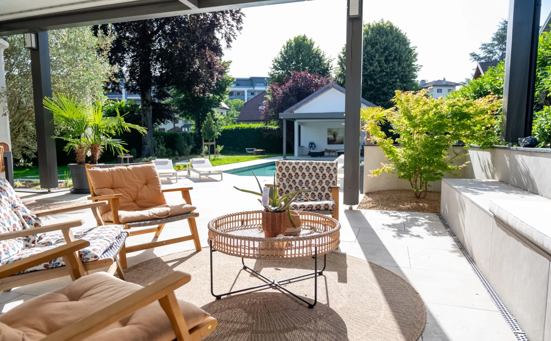 Fauteuils et table basse sous pergola, piscine et pool house en fond – Rumilly